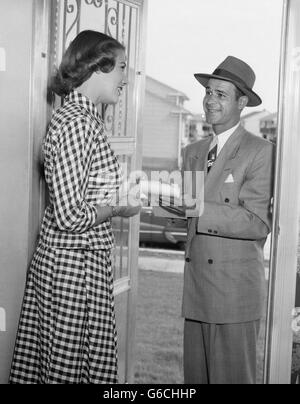 1950ER JAHRE HAUSFRAU IN KARIERTE KLEID AN TÜR, VERKÄUFER IM ANZUG & HUT MIT BLEISTIFT & PAD ZU HAUSE SPRECHEN Stockfoto