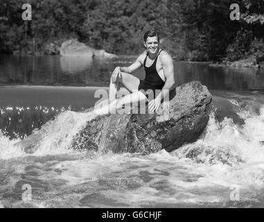 1930ER JAHREN LÄCHELT MAN LOOKING AT KAMERA TRAGEN BADEANZUG TANKTOP BADEHOSE POSIERT AUF GROßE FELSEN IM WALD TEICH SITZEN Stockfoto