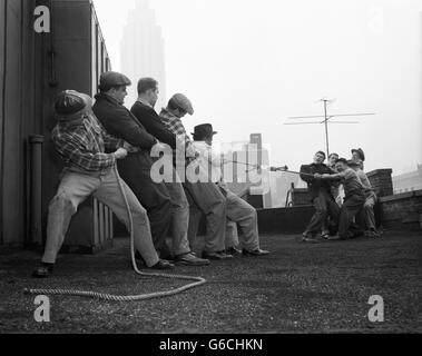 DER 1950ER JAHRE ZWEI GRUPPEN VON FÜNF MÄNNERN ZIEHEN AM TAUZIEHEN SEIL AUF GEBÄUDE DACH VERSUCHEN, ETWAS AUSEINANDER ZIEHEN Stockfoto