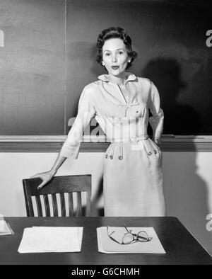 1950ER JAHREN FRAU LEHRERIN BLICK IN DIE KAMERA STAND VOR DER TAFEL IM KLASSENZIMMER STÜTZTE SICH AUF STUHL VOM SCHREIBTISCH Stockfoto