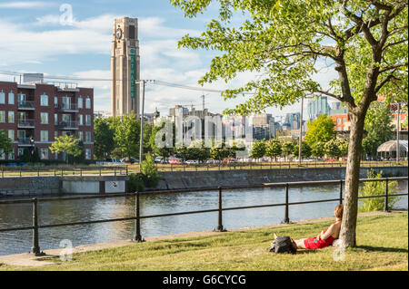 Frau lehnte sich gegen Baum entlang der Lachine Canal in Montreal, Kanada, mit Atwater Market Tower im Hintergrund Stockfoto