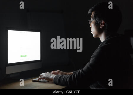 Ernster junger Mann sitzen und arbeiten mit Computer im dunklen Büro Stockfoto