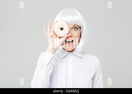 Fröhliche junge Frau in blonde Perücke bedeckt ihr Auge mit Donut auf weißem Hintergrund Stockfoto