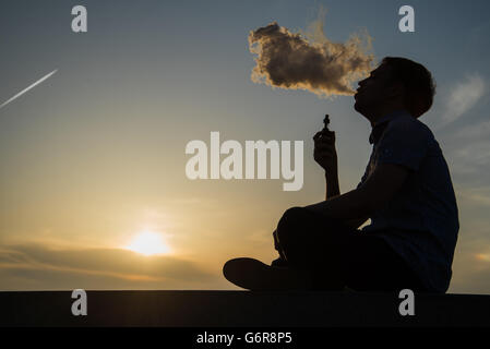 dampfen junger Mann mit, produziert Dampf auf Sonnenuntergang Himmelshintergrund an der Küste Strandpromenade, Platz für text Stockfoto