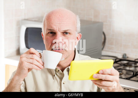 Betreffenden gut aussehend bärtigen reifer Mann mit Glatze und geknöpften Hemd mit gelben Digitalgerät und Kaffeetasse während se Stockfoto