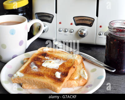 Zwei Scheiben gebuttertes weißes geröstetes Brot auf einem Teller, eine Tasse Tee und ein Glas Marmelade und ein Glas Marmite. Stockfoto