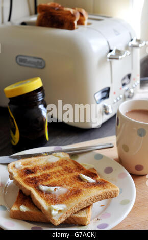 Zwei Scheiben gebuttertes weißes geröstetes Brot auf einem Teller, mit zwei weiteren Scheiben weißen Toast in einem Toaster, einer Tasse Tee und einem Glas Marmelade und Marmite. Stockfoto