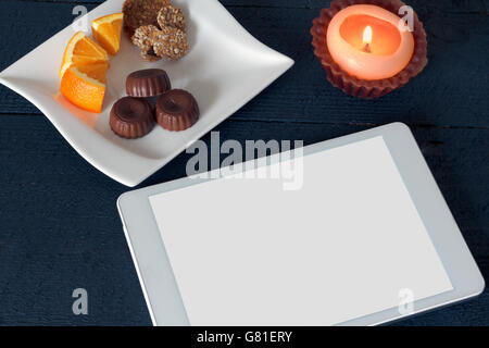 Weiße Tablette auf hölzernen strukturierten schwarzen Hintergrund mit einer Kerze und Süßigkeiten entlang, Raum für Kopie Stockfoto
