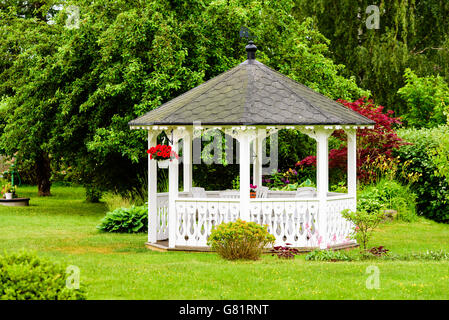 Schöne weiße Pavillon mit roten Blüten, die aus einem Korb hängen. Bäume und Sträucher im Hintergrund. Schönen Garten mit Rasen. Stockfoto