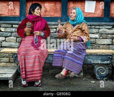 Zwei bhutanische Frauen in traditioneller Kleidung sitzen auf einer Bank und spinnen Gebetsmühle vor einem Steingebäude, um einen friedlichen Moment miteinander zu verbringen Stockfoto