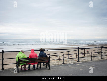 Rückansicht der drei Reife Frauen sitzen auf der Bank mit Blick auf Strand, an einem grauen, kalten Tag in England. UK Stockfoto