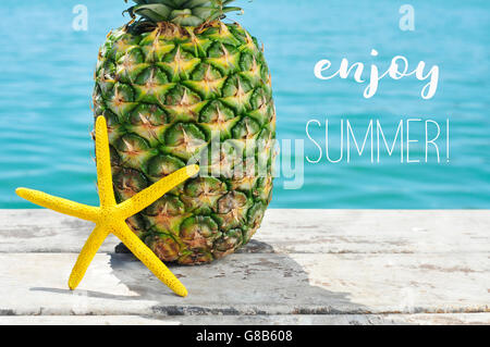 Nahaufnahme von einem gelben Seestern und eine Ananas auf einem verwitterten hölzernen Pier mit dem Meer im Hintergrund und der Text genießen summ Stockfoto