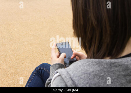 Junge Erwachsene oder späten Teens Frau sitzend, über die Schulter, SMS oder sonst mit ihrem Smartphone gesehen. Fokus liegt auf Telefon. Stockfoto