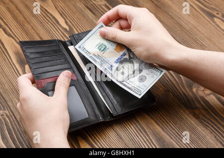 Man setzt 100-Dollar-Banknote in eine schwarze Leder-Geldbörse in dunklem Holz. Schuss von Händen und Brieftasche hautnah Stockfoto