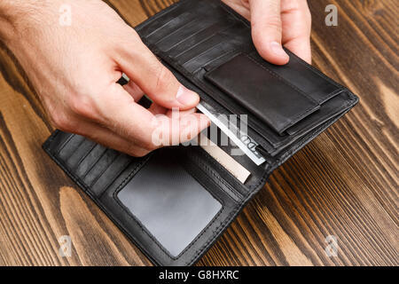 Man setzt 100-Dollar-Banknote in eine schwarze Leder-Geldbörse in dunklem Holz. Schuss von Händen und Brieftasche hautnah Stockfoto