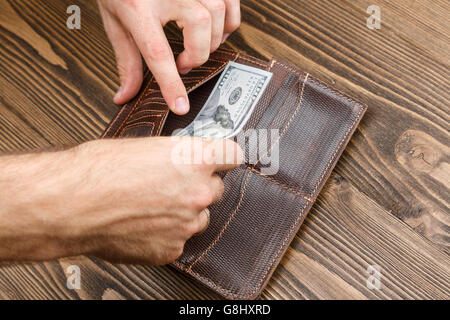 Man setzt 100-Dollar-Banknote in eine braune Leder-Geldbörse in dunklem Holz. Schuss von Händen und Brieftasche hautnah Stockfoto