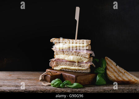 Vollkorn gegrilltes Sandwich-Brot mit schmelzen heiße Käse, Schinken und Basilikum auf Schneidbrett aus Holz in dunklem Hintergrund.