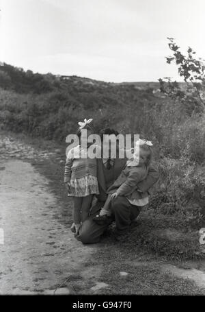 der 1950er Jahre Geschichtsbild, Outdoor-Szene zeigt einen glücklichen Vater kniete hält seine zwei Töchter, England. Stockfoto