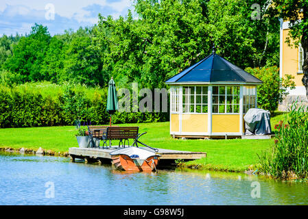 Bjornavad, Schweden – 20. Juni 2016: Kleine orangenes Ruderboot neben einem hölzernen Steg festgemacht. Gelben Pavillon am Rasen hinter sich. Outdoor-f Stockfoto