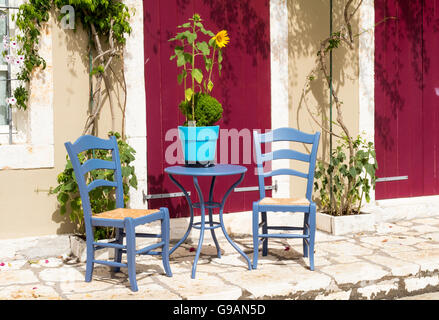 Zwei blaue Bast Stühle, Tisch mit Sonnenblumen Pflanzen in einem Topf mit roten Türen Fiskardo Kefalonia, Griechenland. Stockfoto