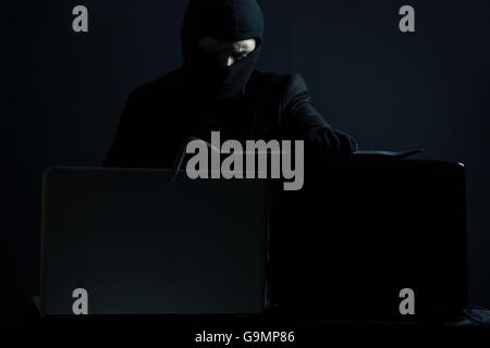 Böse Computerhacker in Anzug stehlen Daten von Laptop mit Brecheisen vor schwarzem Hintergrund Stockfoto