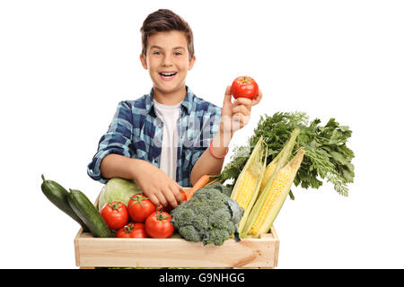 Studio Aufnahme eines fröhlichen jungen posieren hinter eine Kiste voll mit Gemüse und hält eine einzelne Tomate isoliert auf weißem Hintergrund Stockfoto