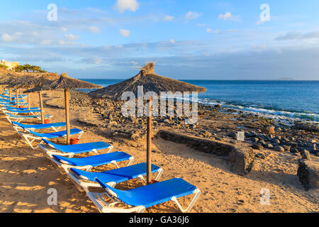 Reihe von Liegestühlen und Sonnenschirmen am Strand von Playa Blanca bei Sonnenuntergang, Lanzarote, Kanarische Inseln, Spanien Stockfoto