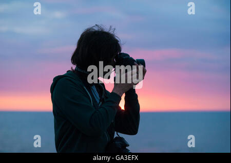 Eine Silhouette eines professionellen Fotografen Mannes mit dem Fotografieren mit einer DSLR-Kamera gegen einen Sonnenuntergang Stockfoto