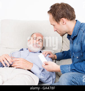 Älterer Mann jüngeren Mann etwas Medizin erhalten, beim liegen auf dem Sofa im Wohnzimmer Stockfoto