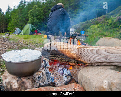 Eine Bratpfanne über einem Lagerfeuer, Wohnmobile und Zelte im Hintergrund. Schottisches Hochland Stockfoto