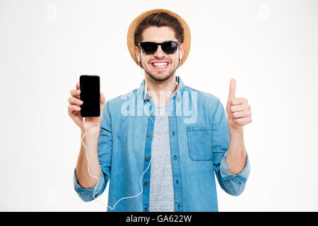 Fröhlichen jungen Mann Kopfhörer zeigen leeren Bildschirm Handy und zeigt Daumen oben auf weißem Hintergrund Stockfoto