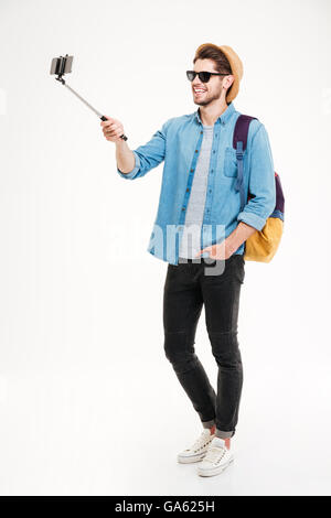 Voller Länge lächelnder jungen Mann mit Rucksack fotografieren mit Smartphone und Selfie stick Stockfoto