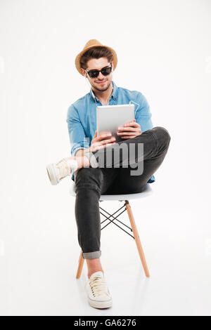 Hübscher attraktive glücklich freudiger junger Mann mit Tablet und sitzen auf dem Stuhl, auf dem weißen Hintergrund isoliert Stockfoto