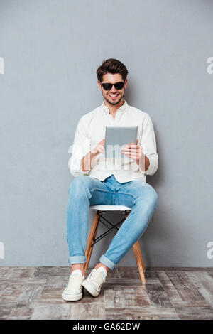 Glücklich Jüngling in Sonnenbrille auf Stuhl sitzend und mit Tablet auf grauem Hintergrund Stockfoto