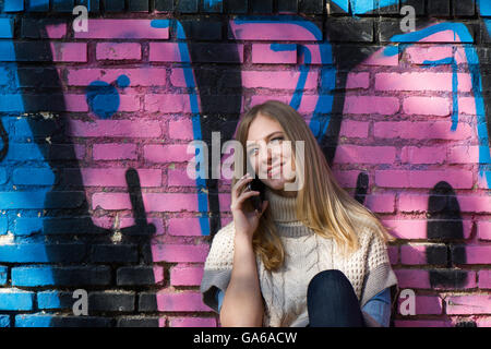 Junge blonde Frau im Gespräch über Telefon und lächelnd beim Sitzen auf dem Boden vor der Ziegelwand mit graffiti Stockfoto