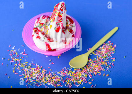 gefrorener Joghurt mit Schwarzkirsche Topping und Regenbogen Streusel auf blauem Hintergrund mit gelber Löffel und Regenbogen-Streusel Stockfoto