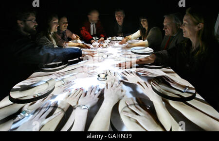 Künstler Terry Flaxton (Mitte rechts) genießt eine virtuelle Mahlzeit mit Gästen in der Winchester Cathedral, einschließlich Rev Canon Roly Riem (Mitte links). Das interaktive Stück mit dem Titel in Other People's Skins zeigt eine Projektion von oben, in der Menschen aus verschiedenen Kulturen zusammen ein Essen genießen. Stockfoto