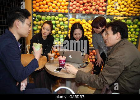 Junge chinesische Männer Saft trinken, reden und schauen einander, eine Frau Freund schaut auf sie, während die beiden anderen auf einem Laptop arbeiten. Stockfoto