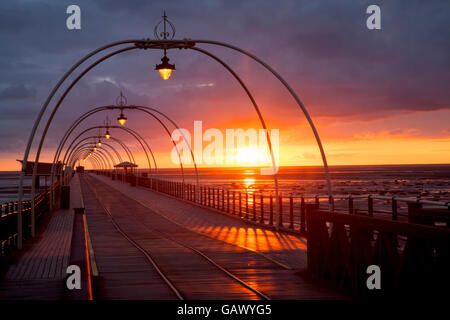 Southport, Merseyside, England. 5. Juli 2016. UK-Wetter: Bunte späten Abend Sonnenuntergang.  Die tief rote Kugel der untergehenden Sonne richtet sich fast mit der Reflexion auf die Straßenbahn-Linien schafft eine Illusion einer Resort-Reise in die Unendlichkeit irgendwo heraus über die irische See Küste, Rötung des nassen Strandes Sand und hölzernen Terrassendielen auf Englands 2. längste Pier.  Bildnachweis: MediaWorldImages/Alamy Live-Nachrichten Stockfoto