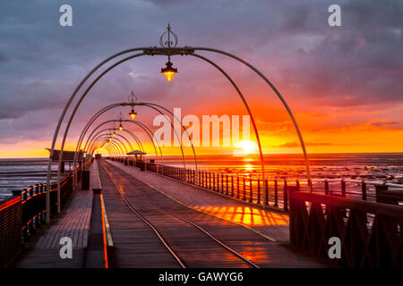 Southport, Merseyside, England. 5. Juli 2016. UK-Wetter: Bunte späten Abend Sonnenuntergang.  Die tief rote Kugel der untergehenden Sonne richtet sich fast mit der Reflexion auf die Straßenbahn-Linien schafft eine Illusion einer Resort-Reise in die Unendlichkeit irgendwo heraus über die irische See Küste, Rötung des nassen Strandes Sand und hölzernen Terrassendielen auf Englands 2. längste Pier.  Bildnachweis: MediaWorldImages/Alamy Live-Nachrichten Stockfoto