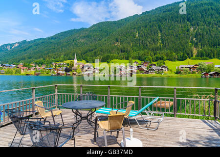 Tisch mit Stühlen auf Holzterrasse und Blick auf grüne Wasser Weißensee im Sommerlandschaft der Alpen, Österreich Stockfoto