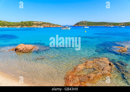 Rote Farbe Felsen am Strand von Santa Giulia mit Booten auf dem azurblauen Meerwasser, Korsika, Frankreich Stockfoto