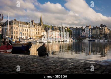 Einsamer Mann sitzt auf der Uferstraße, Blick auf Hafen von Honfleur, Frankreich von der Abendsonne beleuchtet, Frankreich Stockfoto