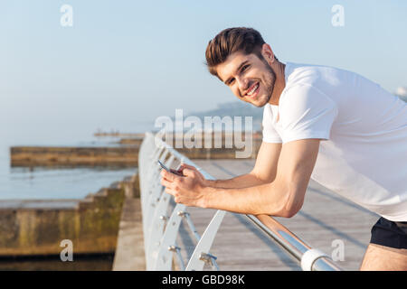 Lächelnde junge Sportler mit Handy stehen auf hölzernen Terrasse am Meer Stockfoto