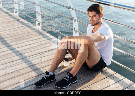 Entspannte junge Sportler mit Flasche Wasser sitzen und entspannen am pier Stockfoto
