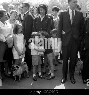 John Kennedy, Sohn des verstorbenen amerikanischen Präsidenten John F. Kennedy, hält sich einen Laib an die Brust und hört zu, wie sein Cousin Anthony Radziwill ihn vor dem Vorplatz des Buckingham Palace in sein Vertrauen nimmt. Sie beobachteten den Wachwechsel, bevor sie die Enten im St. James's Park füttern konnten. Auf sie blicken Jacqueline Kennedy und ihre Tochter Caroline. Stockfoto