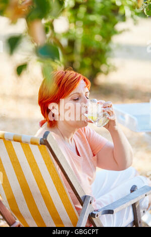 Ältere Frau trinkt ein Glas Wasser auf einem Liegestuhl im Sommer Stockfoto