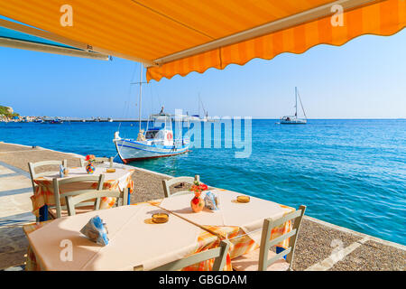 Tisch mit Stühlen im griechischen Taverne auf Insel von Samos mit Fischerboot in Ferne Stadt Kokkari, Griechenland Stockfoto