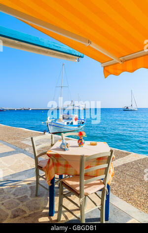 Tisch mit Stühlen im griechischen Taverne auf Insel von Samos mit Fischerboot in Ferne Stadt Kokkari, Griechenland Stockfoto