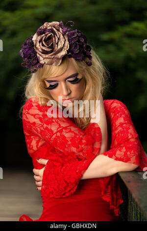 Blonde Modells, feinen Gesichtszügen, purpurrote Blume Krone, geflügelte Eyeliner, riesige Wimpern, rotes Spitzenkleid, kontemplative auf Brücke Stockfoto